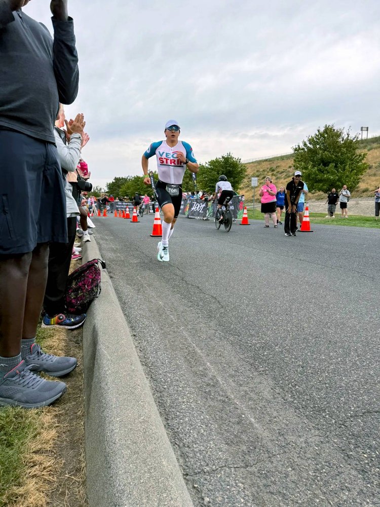 Athlete running during a triathlon, crowds surrounding them on either side. It is clear they are partaking in a triathlon as on the other side of the road there are cyclists riding in the opposite direction.